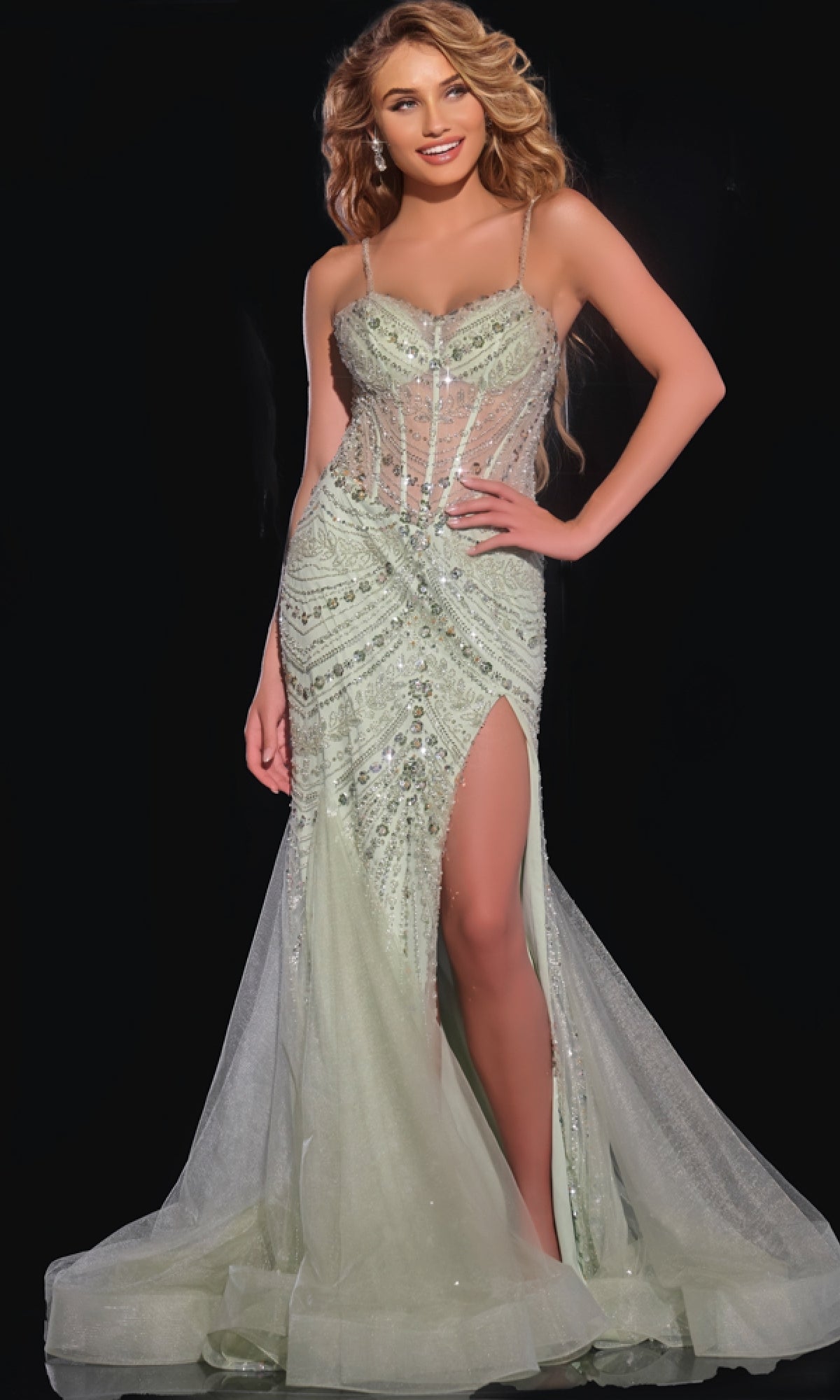 A woman stands against a black background wearing the Jovani 43189 long prom dress—a fitted, light green mermaid gown with sparkly embellishments, sheer corset bodice, and high leg slit.